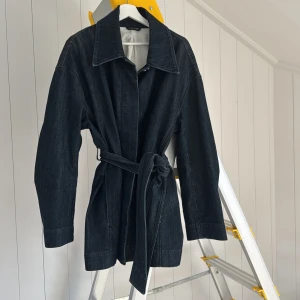 Mörkblå jeansjacka med bälte Filippa K - Snygg mörkblå jeansjacka från Filippa K med klassisk krage, långa ärmar och knytbälte i midjan. Jackan har en rak och lite oversized passform, knappar framtill och är tillverkad i robust denim. Perfekt för dig som gillar stilren och avslappnad look.