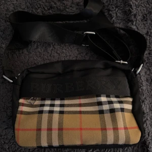 Burberry rutig axelväska med logoband - Säljer en Burberry axelväska med klassiskt rutigt mönster i beige, svart, vitt och rött. Väskan har svart ovandel och baksida, justerbart axelband med Burberry-logga och silverfärgade detaljer. Perfekt storlek för det viktigaste och riktigt snygg streetstil.