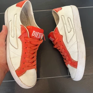 Vita och röda sneakers från Diesel - Snygga sneakers från Diesel i vitt och rött skinn med coola detaljer och tydlig logga på plösen. Skorna har snörning och en klassisk låg profil, perfekt för dig som gillar streetstyle och vill sticka ut med färg. Runda tår och platt sula för skön känsla hela dagen.