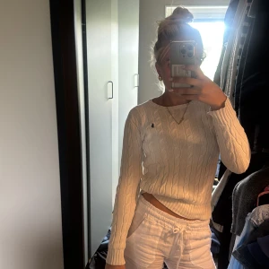 Vit kabelstickad tröja från Ralph Lauren - Vit kabelstickad tröja från Ralph Lauren med klassisk marinblå logga på bröstet. Tröjan har rund halsringning, ribbade muddar och lång ärm. Perfekt för lager-på-lager och snygg till både jeans och kjol.