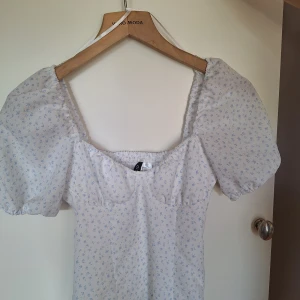 Vit blommig kortklänning H&M XXS - Supersöt vit kortklänning från H&M med ljusblått blommönster. Klänningen har puffiga korta ärmar, smockad rygg och fyrkantig urringning. Perfekt för sommarens varma dagar och ger en romantisk vibe. Endast använd en gång.