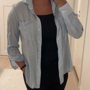 Tunn blus - Snygg blåvit randig blu med klassisk krage och långa ärmar. Skjortan har två bröstfickor och stängs med vita knappar framtill. Perfekt att bära öppen över ett linne eller stängd för en mer uppklädd look. den är helt by och aldrig använd
