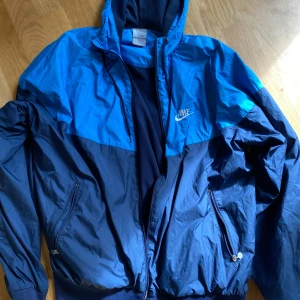 Nike blå vindjacka med huva - Nike vindjacka i två nyanser av blått med broderad logga på bröstet. Jackan har huva med snörning, dragkedja framtill och två fickor med dragkedja. Tillverkad i lätt polyester och har ribbade muddar nertill och vid ärmslut. Perfekt för en sportig look.