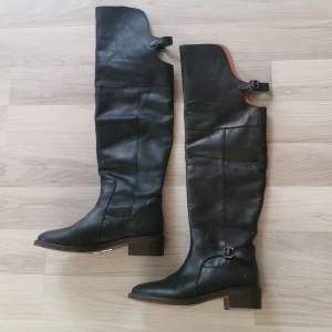 Svarta höga skinnstövlar stl 40 - Snygga svarta stövlar i äkta skinn med högt skaft och låg klack. Stövlarna har spänndetaljer upptill och vid ankeln samt en robust brun sula. Perfekta för dig som vill ha en stilren och edgy look. Haft dom på mig 3 gånger. 