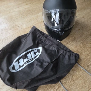 Svart MC-hjälm från HJC - Säljer en svart integralhjälm från HJC med matt finish och klart visir. Hjälmen har ventiler på ovansidan och mjuk vaddering inuti för extra komfort. Medföljer originalpåse med HJC-logga. Perfekt för dig som kör motorcykel och vill ha en stilren look.
