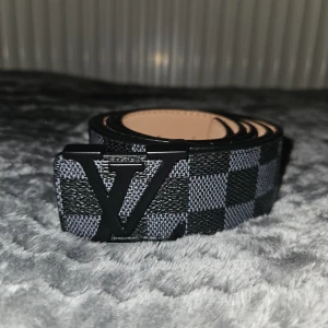 Louis Vuitton rutig svart bälte - Snyggt bälte från Louis Vuitton med klassiskt rutigt mönster i svart och grått. Bältet har en stor LV-logga i svart metall som spänne och är gjort i skinn. Perfekt accessoar för att lyfta din outfit med en lyxig touch.