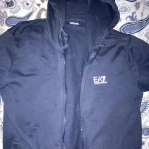 Svart hoodiejacka från EA7 Emporio Armani - Svart hoodiejacka från EA7 Emporio Armani med dragkedja framtill och logga på bröstet. Jackan har huva med snörning och är tillverkad i mjukt bomullsmaterial. Använd endast ett fåtal gånger. Storlek S. 