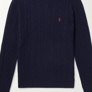 Mörkblå kabelstickad tröja Polo Ralph Lauren - Kabelstickad mörkblå tröja från Polo Ralph Lauren med klassisk röd broderad logga på bröstet. Tröjan har rund halsringning, ribbade muddar och är långärmad. Perfekt för dig som gillar preppy och stilren look.