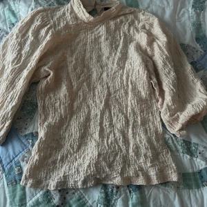 Aprikos blus med struktur från Lindex - Säljer en aprikos blus från Lindex i storlek S. Blusen har långärmade puffärmar, hög krage med knapp i nacken och ett snyggt strukturerat tyg som ger en cool look. Perfekt för dig som gillar stilrena och trendiga plagg.