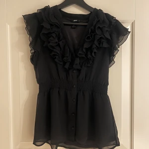 Svart volangblus från H&M - Svart blus från H&M med volanger längs v-ringningen och axlarna. Mycket fint skick!! 