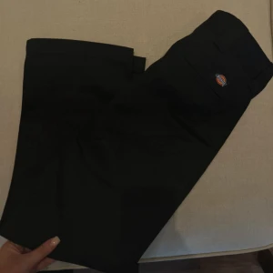 Svarta Dickies byxor med raka ben - Svarta byxor (W28/L30) från Dickies med klassisk rak passform och diskret logga ovanför bakfickan. Byxorna har hällor för bälte och är tillverkade i ett slitstarkt material som känns robust men ändå bekvämt. Perfekta för en clean och stilren look. 