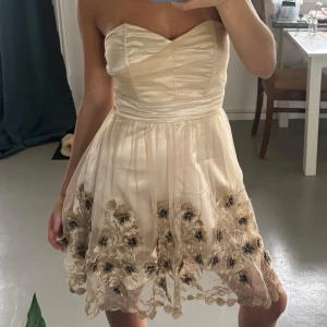 Champagnefärgad axelbandslös klänning - Superfin kort axelbandslös klänning i champagnefärgad satin med voluminös kjol och broderade blommor i guld och svart. Söt hjärtformad urringning och markerad midja. Perfekt för dig som vill sticka ut med eleganta detaljer och romantisk känsla.
