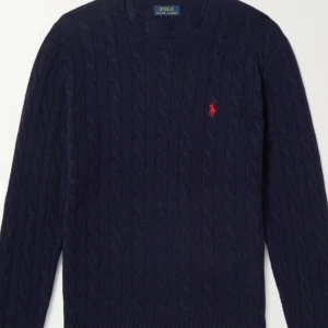 Mörkblå kabelstickad tröja Polo Ralph Lauren - Kabelstickad mörkblå tröja från Polo Ralph Lauren i 100% bomull. Klassisk rund halsringning och lång ärm, med röd broderad logga på bröstet. Perfekt för dig som gillar stilren och tidlös design.