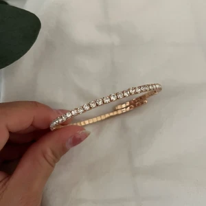 Guldigt armband med glittrande stenar - Superfint armband i guldton med en rad gnistrande klara stenar runt hela bandet. Armbandet är stelt med lätt flexibilitet och har en elegant, tidlös design som verkligen poppar på handleden. Perfekt för dig som gillar att sticka ut med lite extra bling.