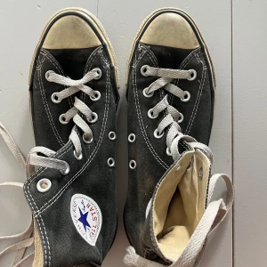 Mörkgråa Converse Chuck Taylor All Star - Klassiska mörkgråa Converse Chuck Taylor All Star med vita snören och rund tå. Skorna har vit sula med svart rand och tydlig logga på sidan. Tillverkade i textil med kontrasterande sömmar och metallöljetter för snörning. Storlek 37,5. 