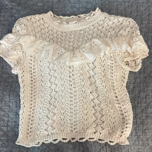 Vit stickad blus med volang från Zara - Supersöt vit stickad blus från Zara med korta ärmar och en bred volang med broderade detaljer över bröstet och axlarna. Blusen har ett romantiskt mönster och ribbad halsringning. Perfekt för dig som gillar en feminin och trendig stil.