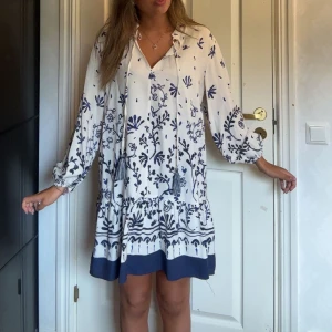 Vit och blå blommig kortklänning - Superfin vit kortklänning från Zara med blått blommigt mönster och volang nertill. Endast använd 1 gång o perfekt till släktträffar och till sommarn