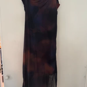 Mörk långklänning med slits - Säljer en långklänning i mörka nyanser med draperad effekt på ena sidan. Klänningen har slits och är ärmlös, perfekt för dig som vill sticka ut. Materialet känns lätt och följsamt.