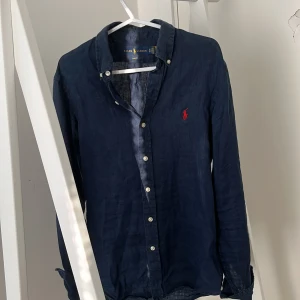 Mörkblå linneskjorta Ralph Lauren XS - Snygg mörkblå skjorta från Ralph Lauren i linne, slim fit-modell med klassisk krage och vita knappar. Ikonisk röd broderad logga på bröstet. Perfekt för dig som gillar stilrena och tidlösa plagg med premiumkänsla. Skick är 7/10 säljer på grund av för liten. Pris kan diskuteras.