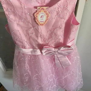 Rosa kortklänning med rosett - Supersöt rosa kortklänning med glittriga broderier och en stor rosett i midjan. Klänningen är ärmlös och har ett tunt lager tyll över kjolen som ger extra volym. Perfekt för speciella tillfällen när du vill känna dig extra fin.