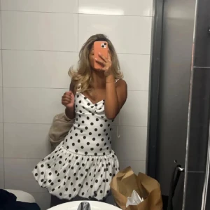 Vit prickig kortklänning med volang - Supersöt vit kortklänning med svarta prickar och volangig kjol. Klänningen har smala axelband och en snygg draperad urringning. Perfekt för dig som vill sticka ut med en lekfull och trendig look.