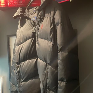 Svart dunjacka Polo Ralph Lauren  - Svart dunjacka från Polo Ralph Lauren med röd broderad logga på bröstet. Jackan har dragkedjor på fickorna och framtill, hög krage och klassisk pufferstil. Perfekt för kalla dagar och riktigt snygg till streetwear. Lite stor i storleken