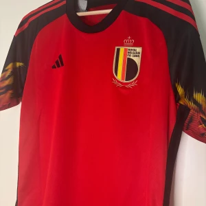 Belgien matchtröja Adidas röd/svart - Snygg Belgien fotbollströja från Adidas i rött och svart med gula detaljer på ärmarna. Använd 1 gång och har ett väldigt skönt material