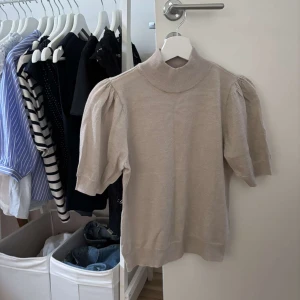 Beige glittrig stickad topp med puffärm från MQ - Snygg beige glittrig stickad topp med kort puffärm och hög krage. Materialet har en lätt glansig finish som ger en lyxig känsla. Perfekt för dig som gillar en stilren look med det lilla extra. Passformen är normal och toppen är enkel att matcha till olika outfits. Märket är Stockh LM och är köpt i MQ