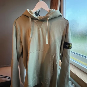 Grön hoodie från Stone Island - Snygg grön hoodie från Stone Island med klassisk patch på vänster ärm. Tröjan har huva med snörning, känguruficka framtill och långa ärmar. Använt ett fåtal gånger. Storlek M