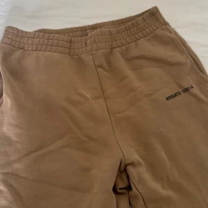 Beige mjukisbyxor från Arigato - Snygga beige mjukisbyxor från Arigato med elastisk midja och diskret svart tryck på ena benet. Byxorna har raka ben och är tillverkade i mjukt bomullsmaterial, perfekta för en chill och avslappnad stil.