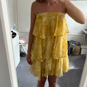 Gul volangklänning NAKD - Superfin kort gul klänning med volanger i flera lager. Sitter snyggt både med och utan axelband som lätt går att vika ner eller sprätta bort. Rosetten i ryggen styr hur långa banden är. Använd en gång på midsommar💛