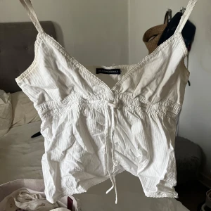 Vit linnetopp från Brandy Melville - Supersöt vit linnetopp från Brandy Melville med smala axelband, v-ringning och knyt framtill. Toppen har en lätt rynkad midja och är croppad, perfekt för varma dagar. Tunt bomullstyg som känns luftigt och fräscht.