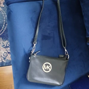 Svart axelväska från Michael Kors - Snygg svart axelväska från Michael Kors i skinn med justerbar rem och stort MK-emblem i guld framtill. Väskan har dragkedja upptill och ett rymligt huvudfack, perfekt för det viktigaste du behöver ha med dig:) Enkel och stilren design som passar till många outfits✨️