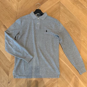 Ralph Lauren piké - Klassisk grå långärmad pikétröja från Polo Ralph Lauren med broderad logga på bröstet. Tröjan har krage, två knappar framtill och är tillverkad i mjuk bomull. Perfekt för en clean och stilren look. Pris kan diskuteras