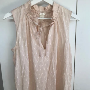 Ljusrosa blus med volang och knyt - Supersöt ljusrosa blus med små vita prickar, volangdetalj vid kragen och knyt framtill. Ärmlös modell i tunn, luftig bomull som passar perfekt till varma dagar. Blusen har en avslappnad passform och v-ringning som ger en chill vibe.