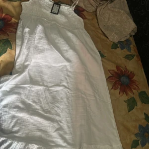 Vit långklänning Ellia med smock - Superfin vit långklänning från Ellia med smock upptill och tunna axelband. Klänningen har en luftig känsla och är perfekt för varma dagar. Söt spetskant upptill och lätt, flowy material som ger en romantisk vibe.