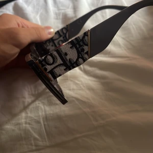 Dior fyrkantiga solglasögon grå - Snygga fyrkantiga solglasögon från Dior med transparent grå båge och mörka glas. Skalmarna har det klassiska Dior-mönstret i svart och vitt, vilket ger en lyxig vibe. Perfekta för dig som vill sticka ut med en statement-accessoar.