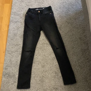 Svarta skinny jeans från LAB Industries - Svarta skinny jeans från LAB Industries i storlek 152. Jeansen har hög midja, klassisk femficksdesign och slitna detaljer vid knäna. Tillverkade i stretchig denim för en bekväm passform. Justerbar midja med knappresår på insidan.