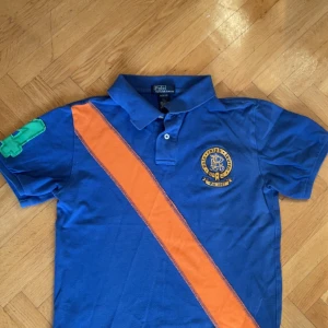 Blå pikétröja Polo Ralph Lauren - Snygg blå pikétröja från Polo Ralph Lauren med orange diagonal rand och broderad logga på bröstet. Tröjan har klassisk krage, korta ärmar och en grön siffra på ärmen. Tillverkad i 100% bomull för en skön känsla. Storlek 8/10!!! 