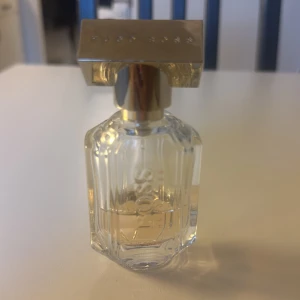 Hugo Boss The Scent parfym - Elegant parfymflaska från Hugo Boss The Scent med genomskinligt, fasetterat glas och lyxig guldig metallkork. Flaskan har en stilren, rektangulär form och innehållet är svagt bärnstensfärgat. Perfekt för dig som gillar exklusiva dofter och snygg design.