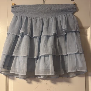 Ljusblå volangkjol med resårmidja - Supersöt ljusblå kortkjol med flera lager volanger och bred smockad resår i midjan. Kjolen är luftig och har en flowy känsla, perfekt för varma dagar. Materialet känns lätt och ger en romantisk vibe.