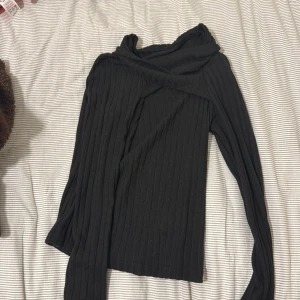 Svart ribbad långärmad tröja - Svart ribbad tröja med lång ärm och snygg omlott-detalj vid halsen. Tröjan är  perfekt för dig som gillar stilrena basplagg med en twist. Materialet känns mjukt och stretchigt, vilket gör den skön att bära.