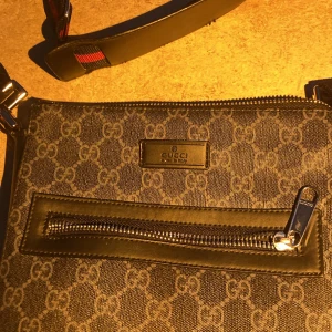 Väska med gg mönster - Säljer en snygg axelväska från Gucci i mörkgrått tyg med klassiskt GG-mönster. Väskan har svarta skinn-detaljer, dragkedja framtill och justerbar axelrem med röd-blå rand. Perfekt för dig som vill ha en stilren och ikonisk väska.