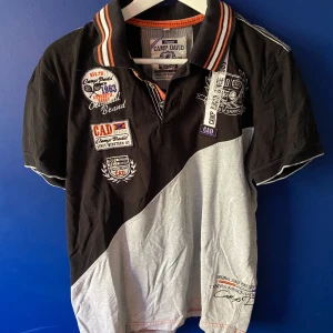 Camp David svart/grå pikétröja - Snygg pikétröja från Camp David i svart och ljusgrått med broderade märken och tryck både fram och bak. Tröjan har korta ärmar, krage med orange och vita ränder samt flera coola detaljer och broderier. Perfekt för dig som gillar sportig och unik stil.