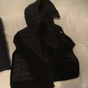 Svart Moncler pufferjacka med huva - Säljer en svart pufferjacka från Moncler med stickade ärmar och huva. Jackan har quiltade partier på kroppen, dragkedja framtill och ribbade muddar. Perfekt för kyliga dagar när du vill ha både stil och värme.