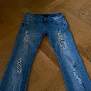 lågmidjade jeans med revor - Snygga blå jeans  med slitna detaljer och hål på både fram- och baksida. Modellen har normal passform, klassisk femficksdesign och dubbla knappar i midjan. Perfekta för dig som gillar en edgy streetstil. Jeansen är bootcut.