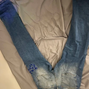Blå slitna jeans med blå tryck - Säljer ett par blå jeans med slitningar och trasiga detaljer på benen. Jeansen har coola blå tryck på låret och stänk av blå färg för en unik look. Klassisk femficksmodell med raka ben och normal passform. Perfekta för dig som gillar streetstyle. Köpta från företag.