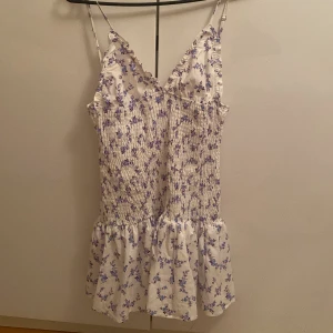 Vit blommig kortklänning med volang - Supersöt vit kortklänning med lila blommor, smockad överdel och volang nertill. Klänningen har smala justerbara axelband och v-ringad hals med små volangdetaljer. Perfekt för varma dagar och somriga vibes!