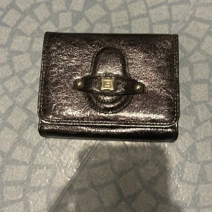 Svart plånbok från Laura Biagiotti - Snygg svart plånbok från Laura Biagiotti i glansigt skinn med struktur. Plånboken har flera fack för kort, myntfack med dragkedja och en cool metallogo på framsidan. Perfekt för dig som vill ha stil och ordning på dina grejer.