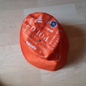 Orange bucket hat med loggor - Cool orange bucket hat med massor av vita loggor från olika märken som Adidas, Polo, Tommy Hilfiger och Levi's. Hatten är i mjukt tyg och har bred brätte runtom. Perfekt för att sticka ut i mängden och ge outfiten en färgklick.